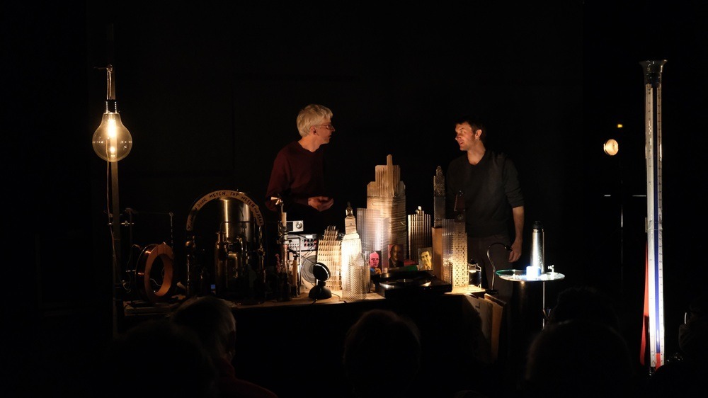 Scène de conférence scientifique sur la supraconductivité : deux chercheurs, Hugues Pothier et un collaborateur, discutent sur une table remplie d’instruments scientifiques et d’objets poétiques, éclairés par des lumières chaudes, devant un public dans l’obscurité. 