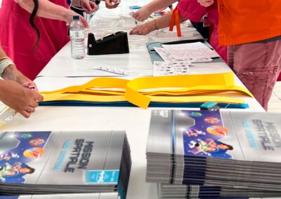 Atelier créatif de la Journée Science à l'hôpital Robert-Debré : bénévoles des Blouses Roses en tabliers roses préparant du matériel pédagogique sur une table. Piles de brochures "Mission Spatiale - Pack Aventure" au premier plan, bandes de papier colorées jaunes et bleues.