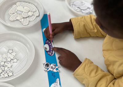 Atelier créatif de la Journée Science : un enfant en veste jaune colorie un ovni violet sur une bande de papier bleue décorée d'astronaute et de planète. Assiettes contenant des découpes en papier (fusées, ovnis, étoiles) à disposition. Activité manuelle pour explorer l'espace.