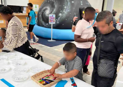 Journée Science à l'hôpital Robert-Debré : le planétarium gonflable au centre du hall, avec des visiteurs de tous âges autour des tables d'ateliers. Un jeune enfant choisit des feutres colorés dans un panier en osier. Ambiance familiale et intergénérationnelle.