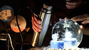 Expérience scientifique en laboratoire : une main tient une bouteille métallique au-dessus d'une boîte émettant de la fumée blanche, sous une lumière bleutée, avec des équipements techniques en arrière-plan.